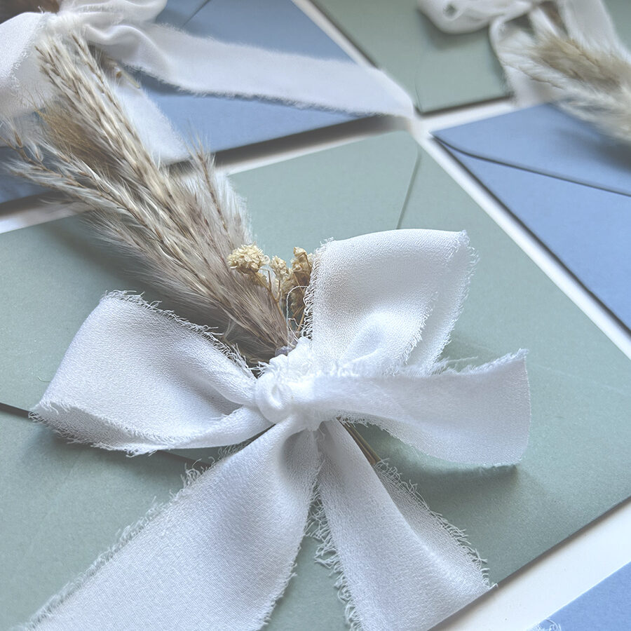 Anteprima del catalogo partecipazioni online 2026 con busta colorata, nastro bianco in chiffon di seta e mini bouquet di fiorellini secchi