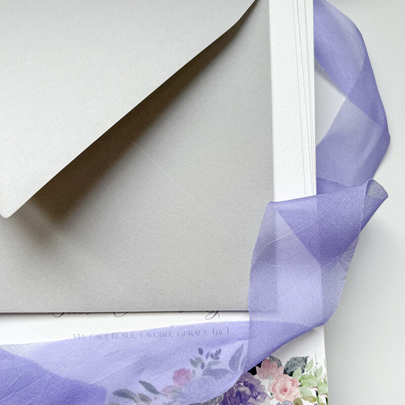 Inviti di matrimonio eleganti con buste grigio perla, carta pregiata e nastri in organza lilla, con dettagli floreali acquerellati realizzati da Cupcake Graphics Studio